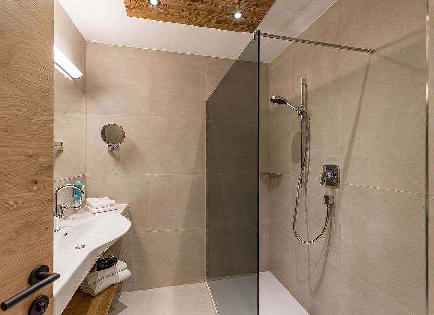 Modernes Badezimmer mit offener Dusche, Glastrennwand, großem Spiegel und Waschbecken auf Holztisch. Warme Beleuchtung verleiht gemütliche Atmosphäre.