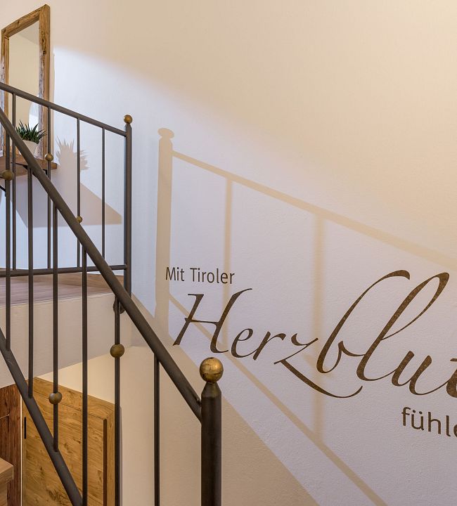 Ein Treppenhaus mit hölzernen Stufen und einem schwarzen Geländer. An der Wand steht: "Mit Tiroler Herzblut fühlen." Dekoration mit Herzmotiv auf Regal.