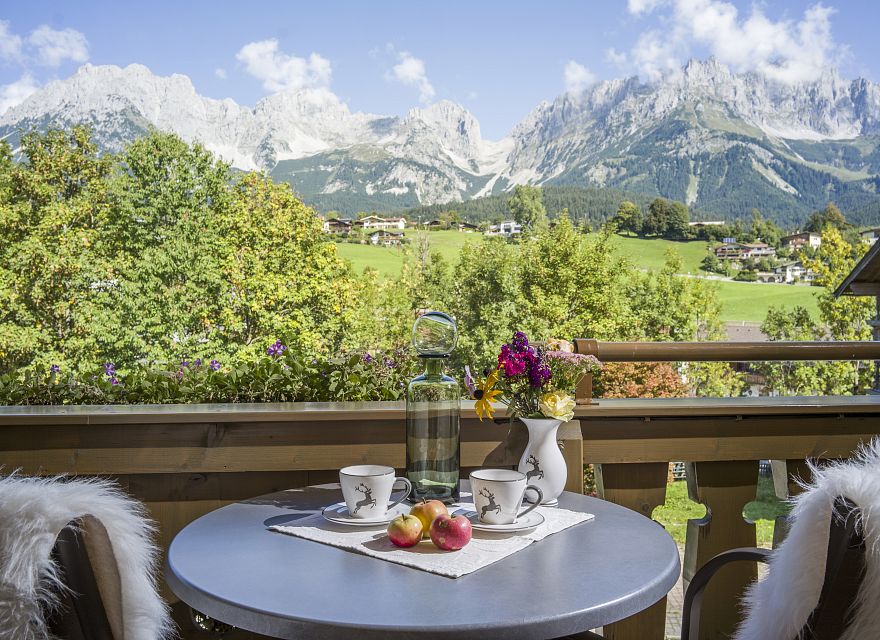 Ein runder Tisch auf einem Balkon bietet Frühstück mit Kaffee, Blumen und Äpfeln. Im Hintergrund sind saftig grüne Wiesen und beeindruckende Berge zu sehen.