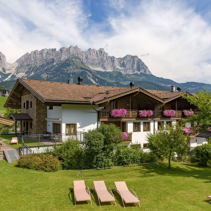 Ein Landhaus mit blühenden Balkonen steht vor einer beeindruckenden Bergkulisse. Der Garten ist gepflegt, mit Sonnenliegen auf dem grünen Rasen verteilt.